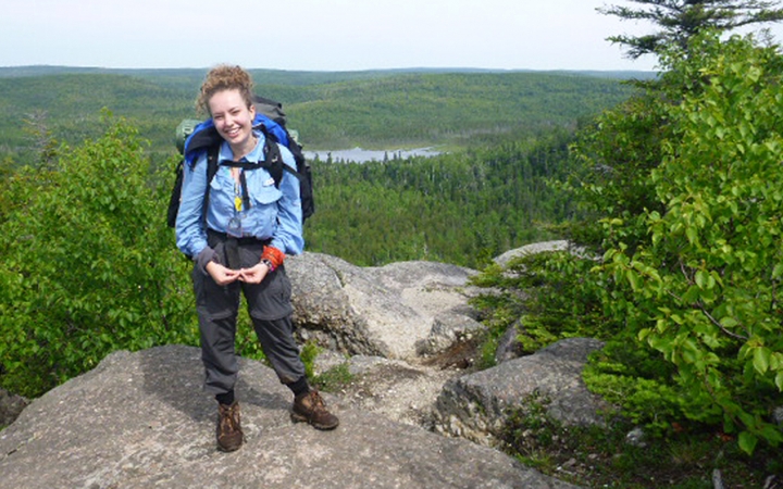 A person wearing a backpack stands on a rocky outlook, smiling at the camera. Behind them stretches a vast green forrest and a body of water. 